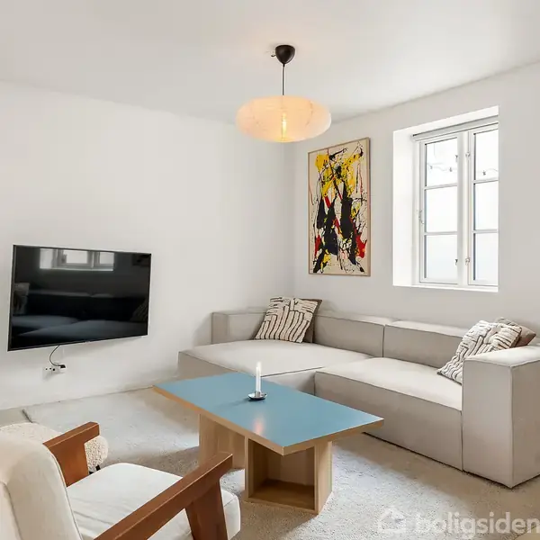 En beige hjørnesofa med puder står i en lys stue. Et moderne maleri hænger på væggen nær et vindue. Der er et fladskærms-TV og et sofabord.
