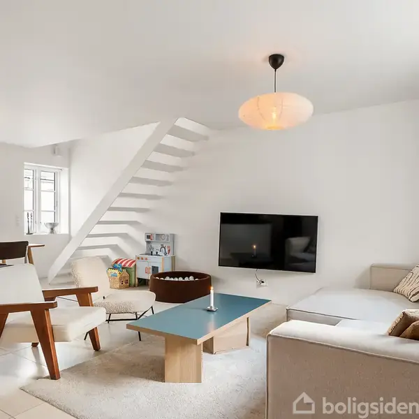 Sofa står i et moderne, lyst opholdsrum. Ved siden af er et sofabord og en lænestol. En fladskærm hænger på væggen. Spisebord og stole ses i baggrunden.