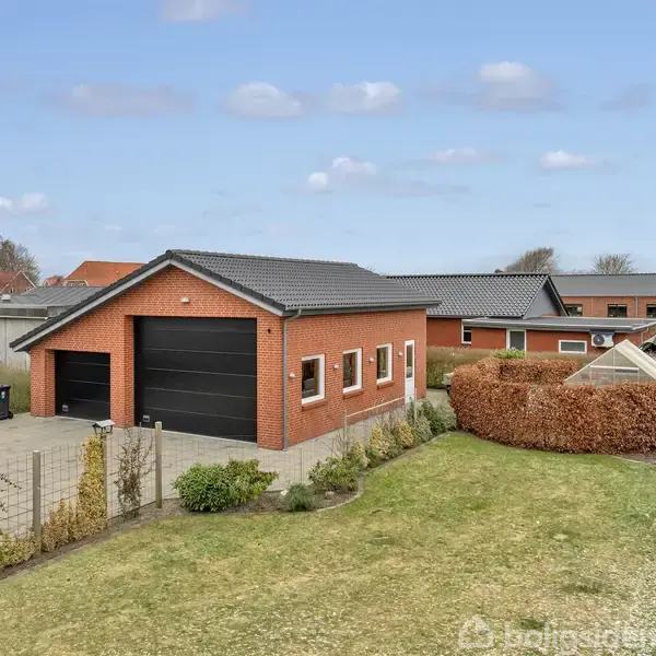 En rød murstensgarage med flere vinduer og en stor sort port står ved en velplejet have med plæne. I baggrunden ses flere bebyggelser og et klart, blåligt himmellandskab.