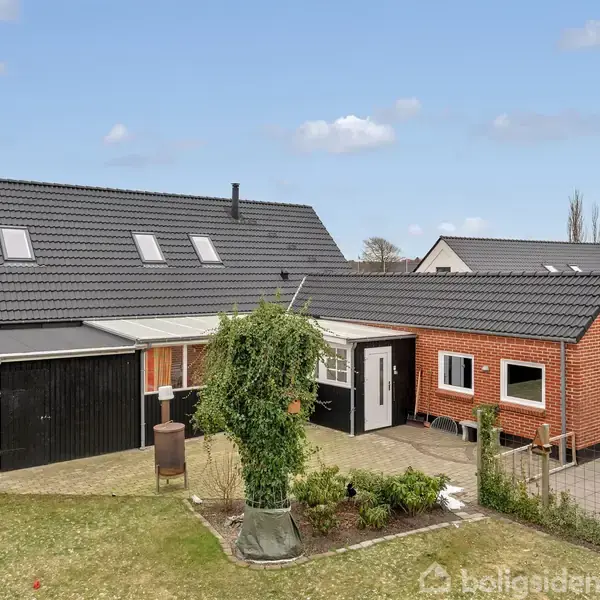 Hus med røde mursten og sort tag med kviste, omgivet af have og flisebelagt indkørsel. Til venstre ses en garage, og der er buske og græs i forgrunden.