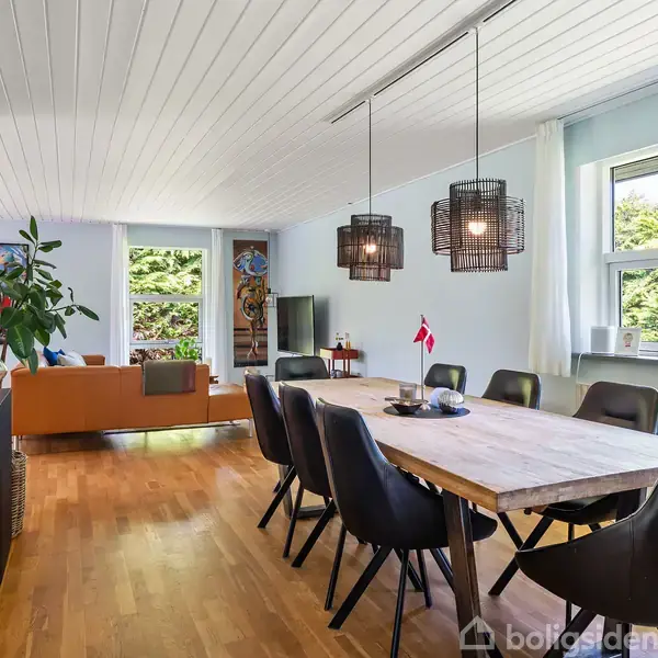 Et langt spisebord med sorte stole står i en stue. Lampen over bordet lyser rummet, der har trægulve, store vinduer, planter og kunstværker. To små dannebrogsflag pynter på bordet.