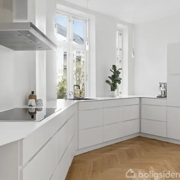 Hvidt køkken med moderne design, kogeplade på venstre side, urtepotteskjuler i hjørnet og flaske på bordet. Trægulv og store vinduer, der giver naturligt lys.