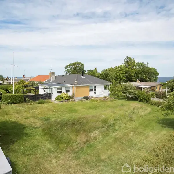 Et enkelt etplanshus står stille i en åben have med højt græs, omgivet af få træer og buske. Baggrund viser flere huse og en udkig til havet under en klar himmel.
