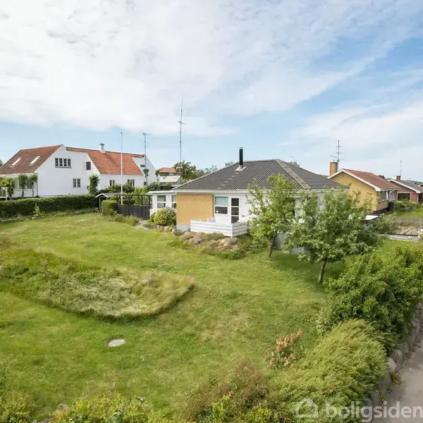 Et gult hus med sort tag står stille ved en vej i et grønt boligkvarter. Det er omgivet af en stor græsplæne og nabolagets huse.