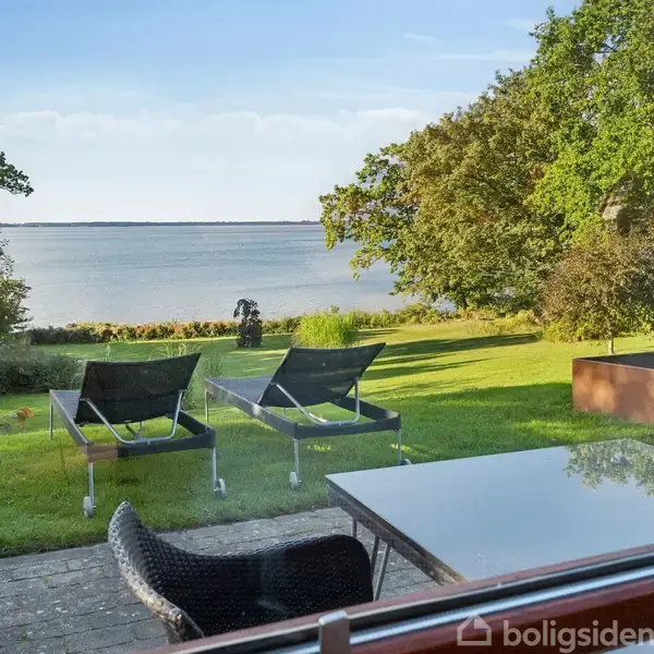 To liggestole står på en velplejet græsplæne med udsigt over en rolig sø. Omkring er der frodige træer og en terrasse med et bord og stol.