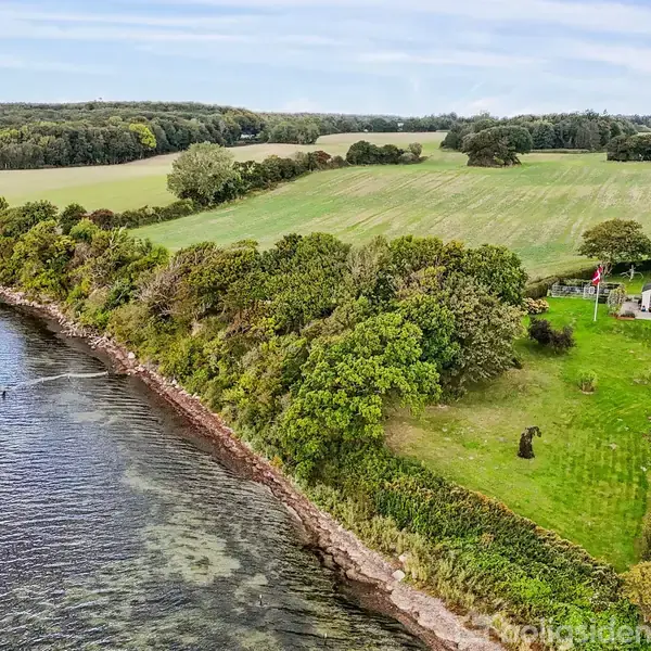 Hus ligger på en grønt græsareal ved siden af en skovbevokset kystlinje. Området omkring huset har marker og spredte træer.