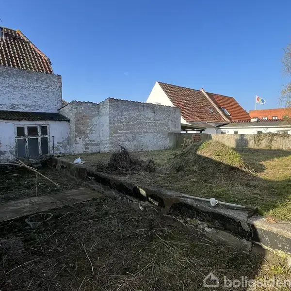 En ubebygget grund, dækket af græs og jord, ligger omgivet af ældre, delvist afskallede hvidmalede mure. I baggrunden ses et par huse med røde tegltage.