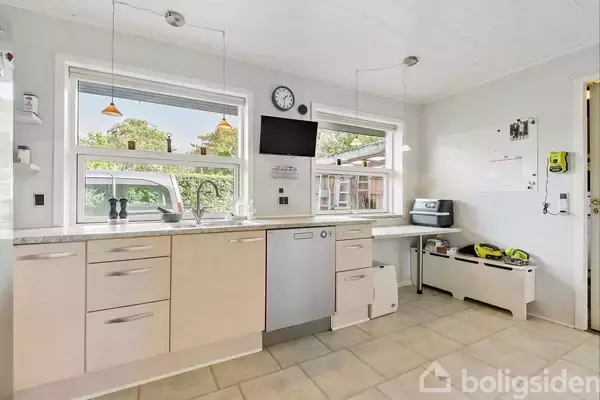 Et lyst køkken med beige skabe og en opvaskemaskine i midten. To store vinduer giver udsigt til træer udenfor. På væggen hænger en sort skærm og et ur.