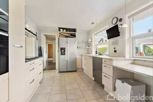 Et moderne køkken med lys indretning, hvor et stort køleskab står mellem skabe. Rummet er lyst med vinduer og har en vægmonteret skærm og ur i hjørnet.