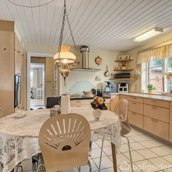 Et køkkenbord med en hvid dug og fire stole står i centrum, omgivet af træskabe og køkkenudstyr. Vinduer med gardiner lader lys strømme ind, og der hænger en lampe over bordet.