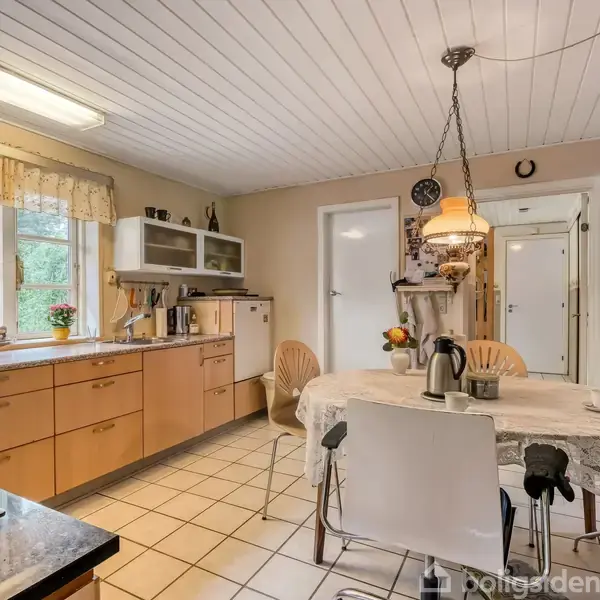 Et køkken med trækøkkenskabe og en bordplade ved vinduet. Et spisebord med en dug, kaffekande og kopper. Omgivelserne inkluderer stoler og udsyn til en grøn have.