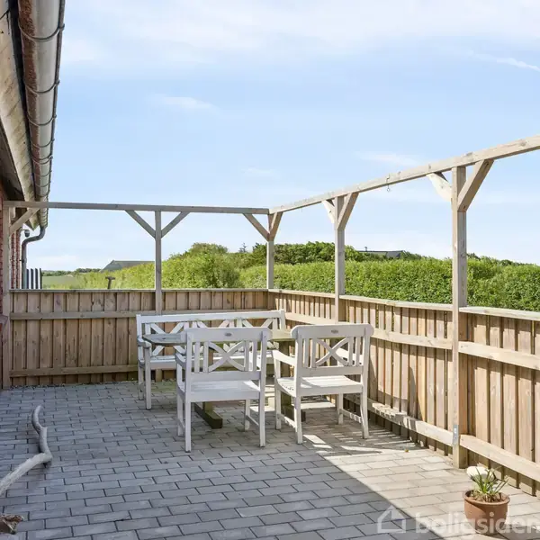 Et stort hus med en grå-hvid facade står midt på en grøn græsplæne. Træer omgiver huset, og der er blå himmel i baggrunden.