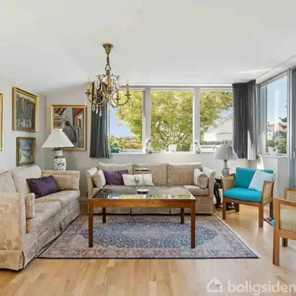 En stue med en beige sofa og lænestole, sofabord, tæppe og flere malerier på væggen. Store vinduer giver udsigt til frodige træer og en have.