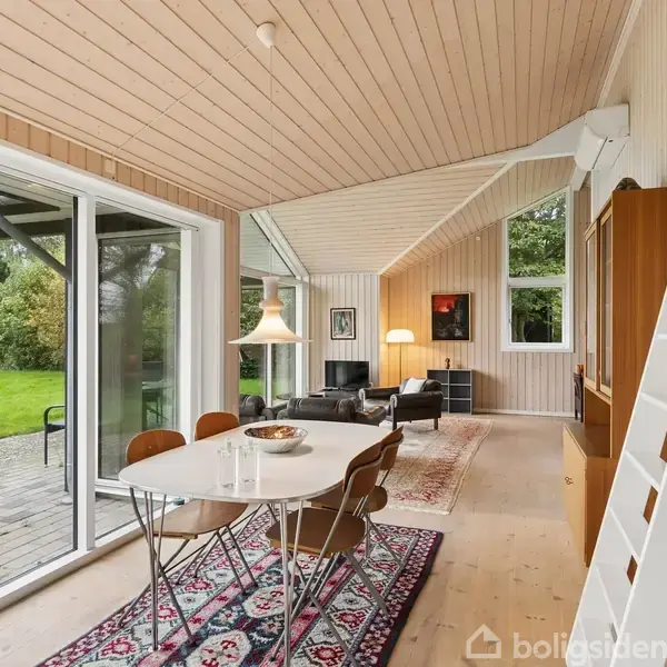 Et spisebord med stole står på et mønstret tæppe i en lys stue. Store vinduer giver udsigt til en grøn have. En stige leder op til en hems.