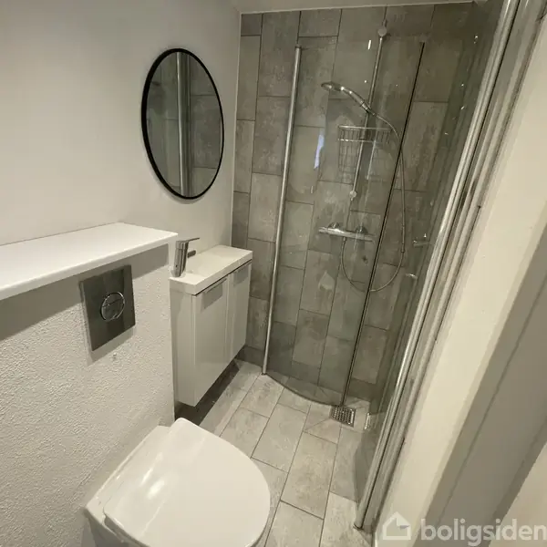 Et moderne badeværelse med brusekabine, væghængt toilet og rundt spejl over en lille håndvask. Fliser dækker gulv og brusevæg, mens resten af rummet er hvidt.