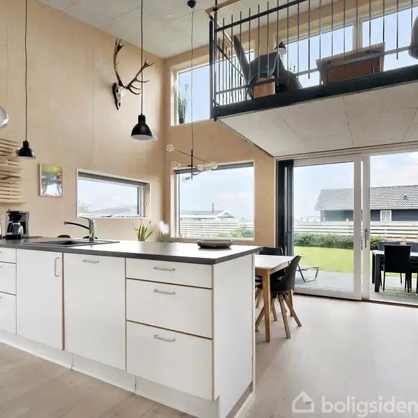 Hvidt, moderne køkken med sort bordplade og ophængte lamper; åbner til spisestue med stort vindue og udsigt til have; ovenpå en hems med gelænder.