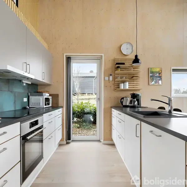 Et moderne køkken med hvide skabe og sorte bordplader. En ovn og mikrobølgeovn er indbygget. En dør leder til en have. Vægur og vindue ses.