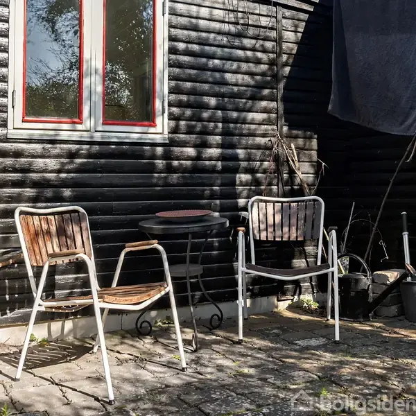 To stole står ved et lille rundt bord på en flisebelagt terrasse foran et sort træhus. Solen skinner på området.