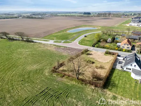 Et luftfoto viser en åben, græsbevokset mark med en enkelt stor træ midt i. Der er en vej og et hus i nærheden, omgivet af landbrugsjord og en lille dam.