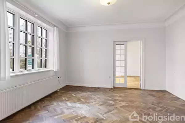 Et tomt værelse med trægulv og store vinduer til venstre. En dør med glasruder fører til et andet værelse. Der hænger en loftslampe.