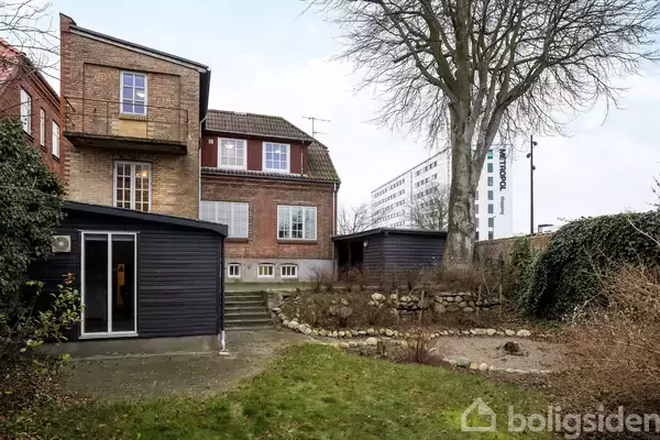 En murstensbygning med to etager står ved siden af en mindre sort tilbygning. Foran er der en have med græs og en stor træbevoksning. I baggrunden ses et moderne, hvidt højhus.