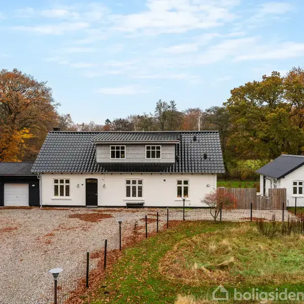 Et hvidt hus med sort tag står stille omgivet af efterårstræer og grønne marker. Ved siden af huset ses en mindre bygning samt en indkørsel af grus.