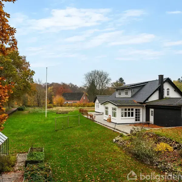 Hvidt hus med sort tag står i en grøn have omgivet af efterårstræer; der er et drivhus og en flagstang nær huset.