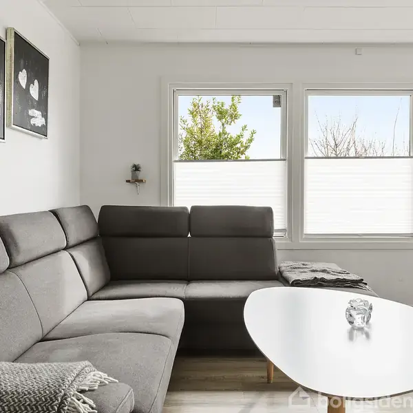 Grå sofa med tæppe ligger op ad væggen, hvorpå hænger to billeder med hjerter. Foran er et hvidt ovalt sofabord med glasdekorationer. Vinduet viser træer udenfor.