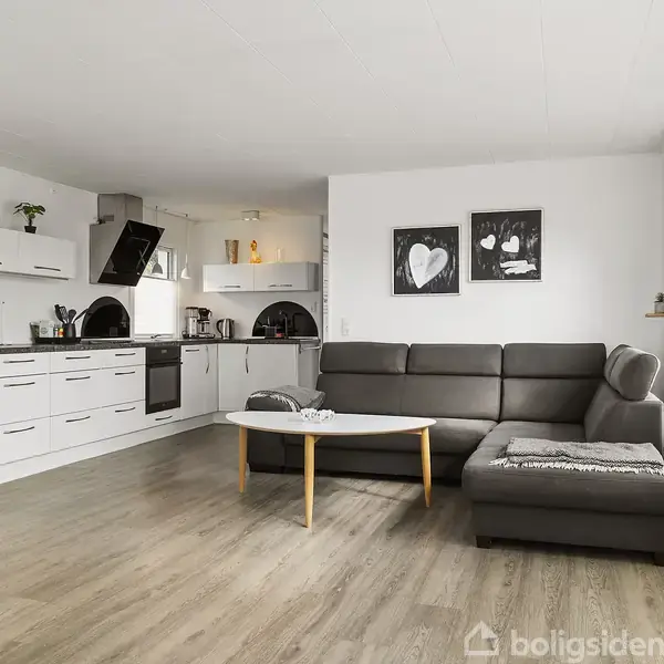 Sofaen står i stuen ved et sofabord, mens et moderne køkken med hvide skabe og apparater er i baggrunden. To billeder hænger på væggen.