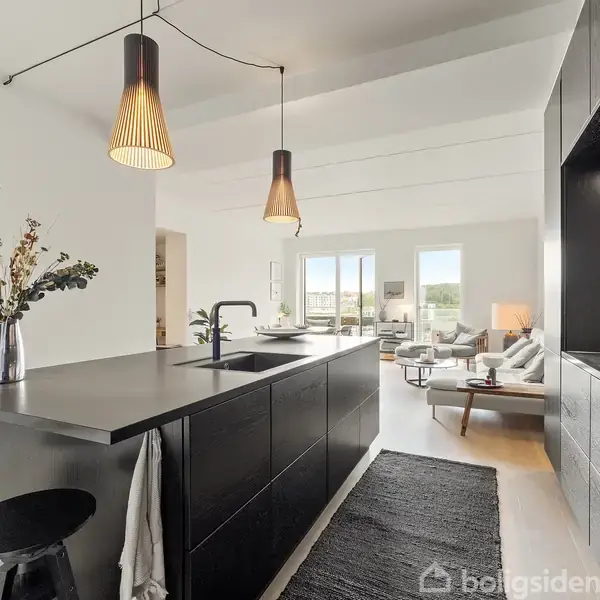 Køkkenø med sort bordplade og to barstole, flankeret af pendellamper. Stuen i baggrunden har store vinduer og lyse møbler. Blomsterdekoration på bordet.