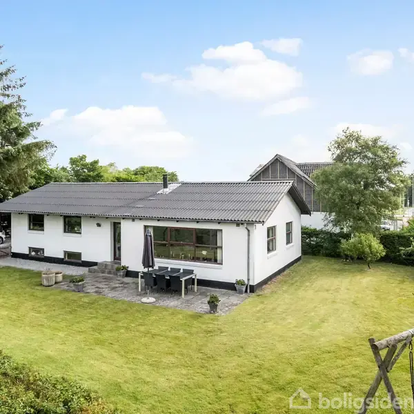 Et hvidt hus med mørkt tag står på en velplejet græsplæne. Foran huset ses en terrasse med havemøbler og parasol. Bilen står parkeret ved siden af huset.