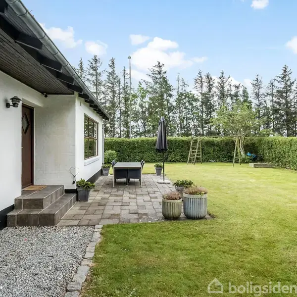 Et hvidt hus har en terrasse med bord og stole. Græsplænen er omkranset af hække, med en vippe og skov i baggrunden. Ingen tekst til stede.