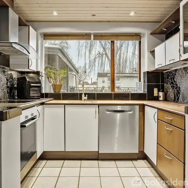 Et moderne køkken med hvide skabe og sorte bordplader. En opvaskemaskine og ovn er indbygget. Vindue med udsigt til en have. Klinkegulv og mosaik backsplash pryder rummet.