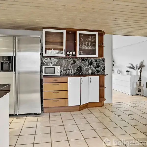 Stål-køleskab står ved siden af et skab med glaslåger og en mikrobølgeovn. Fliser på gulvet og træloft. Åbning fører til opholdsstue med planter og dekorationer.