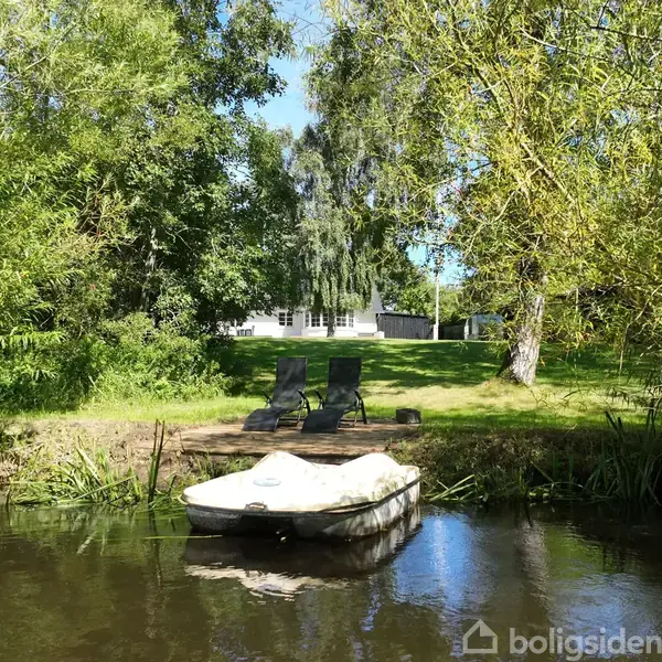 Båd ligger stille i en lille sø omgivet af frodig skov. I baggrunden står to tomme stole på bredden foran et hus, indrammet af grønne træer.