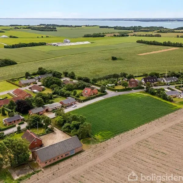 Byområde med røde hustage ligger blandt frodige landbrugsmarker og skovområder. I baggrunden ses havet og små øer mod horisonten under en klar himmel.