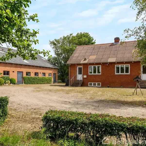 Et rødt murstenshus med hvide vinduer og en skrånende tag, stående på en gårdsplads omgivet af grønne buske og træer. Ved siden af er der en stor garagebygning.