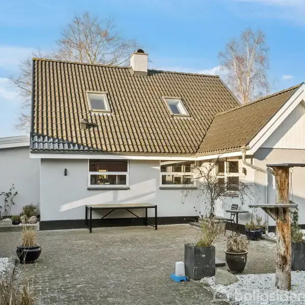 Et hvidt hus med gråt tag står i en gårdhave. Omkring huset er der krukker med planter, en bænk under vinduet og et åbent himmelrum.