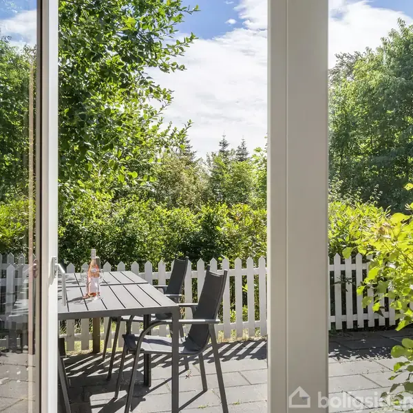 En hvid dør står åben mod en terrasse med et bord og stole. Bordet har en flaske på sig. Haven omkring terrassen er frodig med grønne buske og et hvidt stakit.