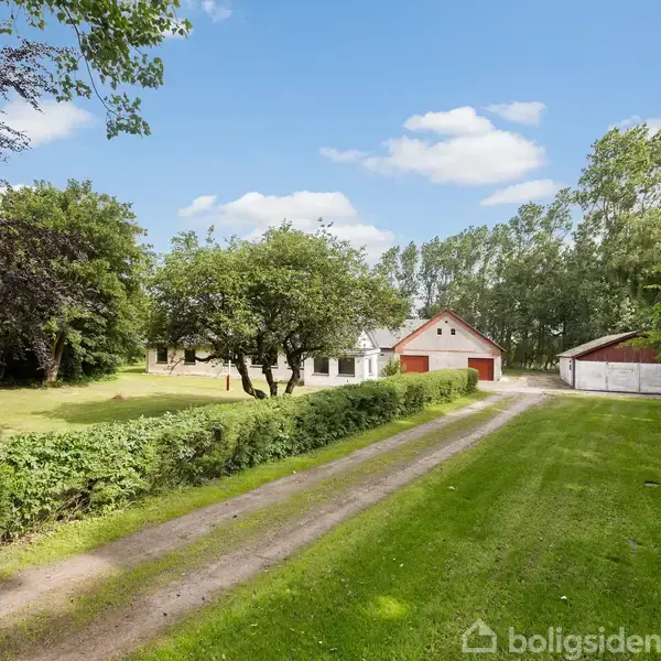Hus i landligt område omgivet af træer og græsplæner. En gruset vej løber gennem omgivelserne, mens himlen er blå med spredte skyer.