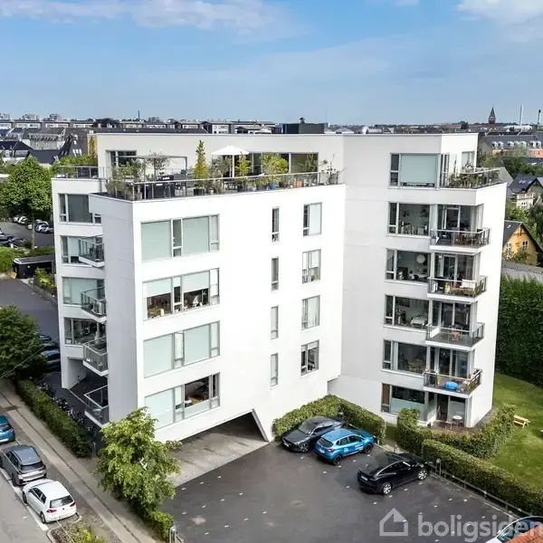 En hvid fem-etagers lejlighedskompleks, omkranset af parkeringspladser og grønne træer, med store vinduer og altaner. Foran er der parkerede biler, og i baggrunden ses byens skyline.