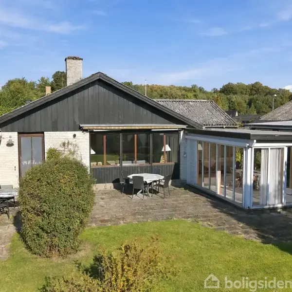 Hus med sort tag og terrasse, omgivet af græsplæne og tæt buskads. Til højre er der en udestue med glaspartier. Himlen er klar og blå.