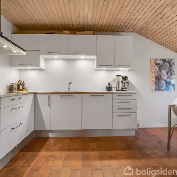 Køkken med hvide skabe og indbygningsovn, der står tomt. Flisebelagt gulv og træbeklædt loft. Et billede hænger på væggen, og et bord med stole står ved vinduet.