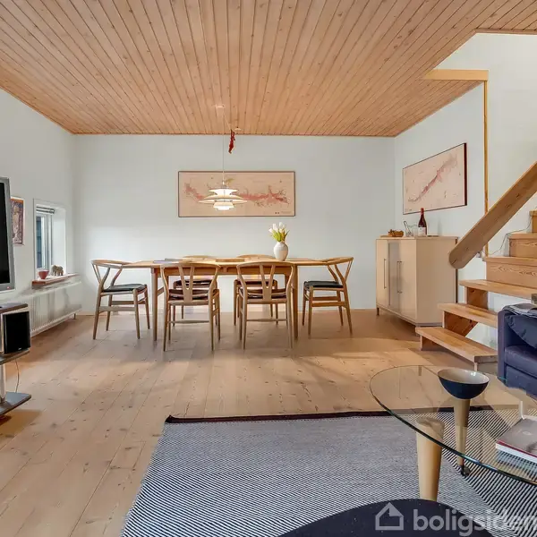 Stue med spisebord og seks stole midt i rummet. Tv står på skænk til venstre; sofa og glasbord til højre. Træloft og trappe i baggrunden. Maleri på væggen.