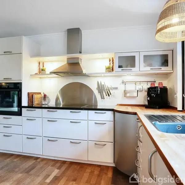 Et moderne køkken med hvide skabe og indbyggede apparater. Der er en ovn, emhætte og sort kaffemaskine på bordpladen. Køkkenet har en stor vask ved vinduet, omgivet af træelementer.