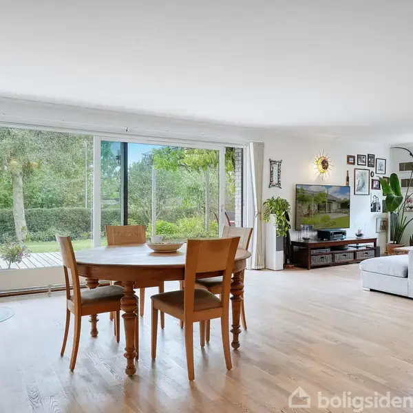 En spisebordsgruppe med fire stole står foran en stor glasskydedør. Udenfor ses en frodig have. I stuen er der en pejs til venstre og en sofa samt et fjernsyn til højre.