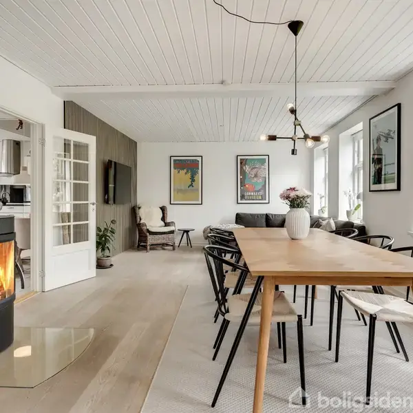 Brændeovn opvarmer stue, hvor et langt spisebord med stole står. Rummet er lyst med hvide vægge og loft. Store vinduer og vægkunst skaber en moderne, hyggelig atmosfære.