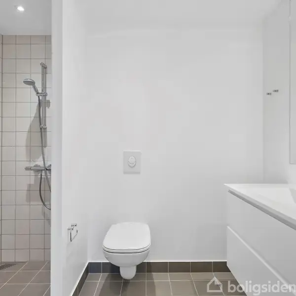 En moderne badeværelse med væghængt toilet, bruser i et flisebelagt område, og en hvid vask med skab. Rummet er lyst med hvide vægge og grå flisegulv.