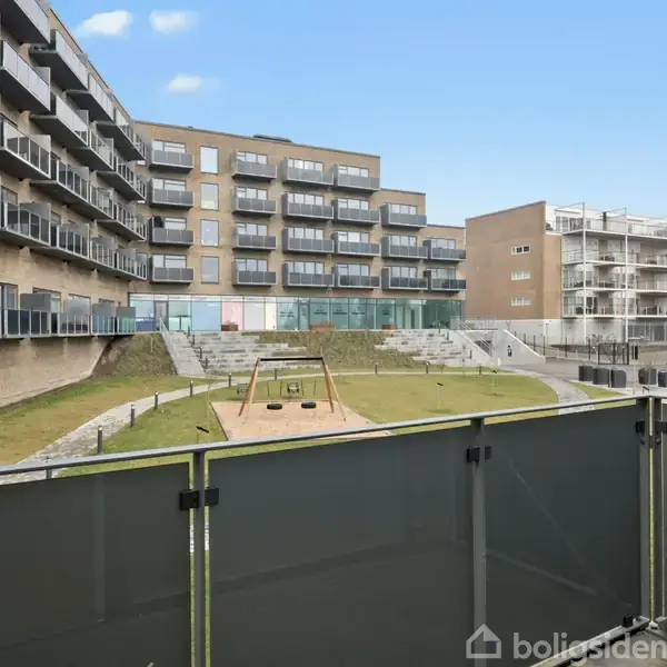 En legeplads med gynger i bevægelse ses fra en balkon, omgivet af moderne boligblokke med altaner. Bygningerne står i solskin under en blå himmel.