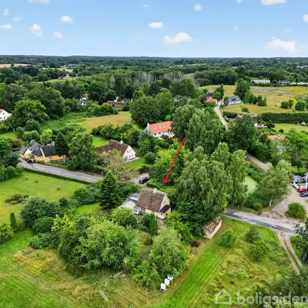 Hus, omgivet af frodig vegetation, står mellem store træer i et landligt område. Området er præget af marker og spredte huse, med en vej foran. Rød pil peger på huset.